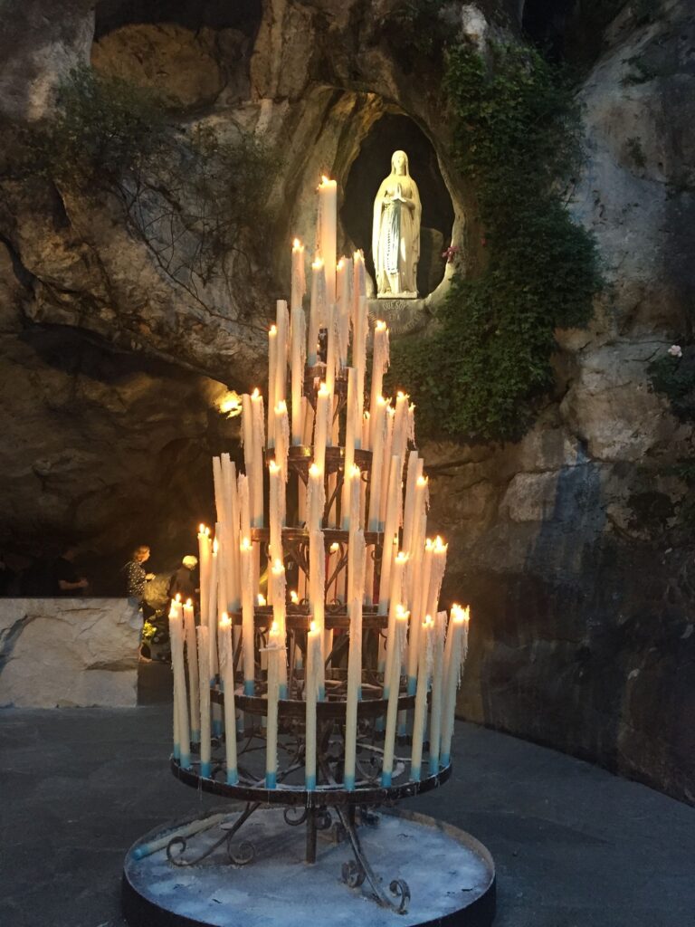 A tall candle stand lit in prayer beneath the statue of the Virgin Mary at Lourdes Grotto at night.
